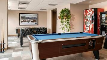 A pool table in a room with a framed picture on the wall, a plant, and a vending machine.