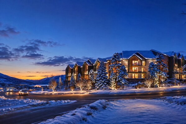 A multi-story lodge with a snow-covered landscape, a road in the foreground, and a mountain range in the background.