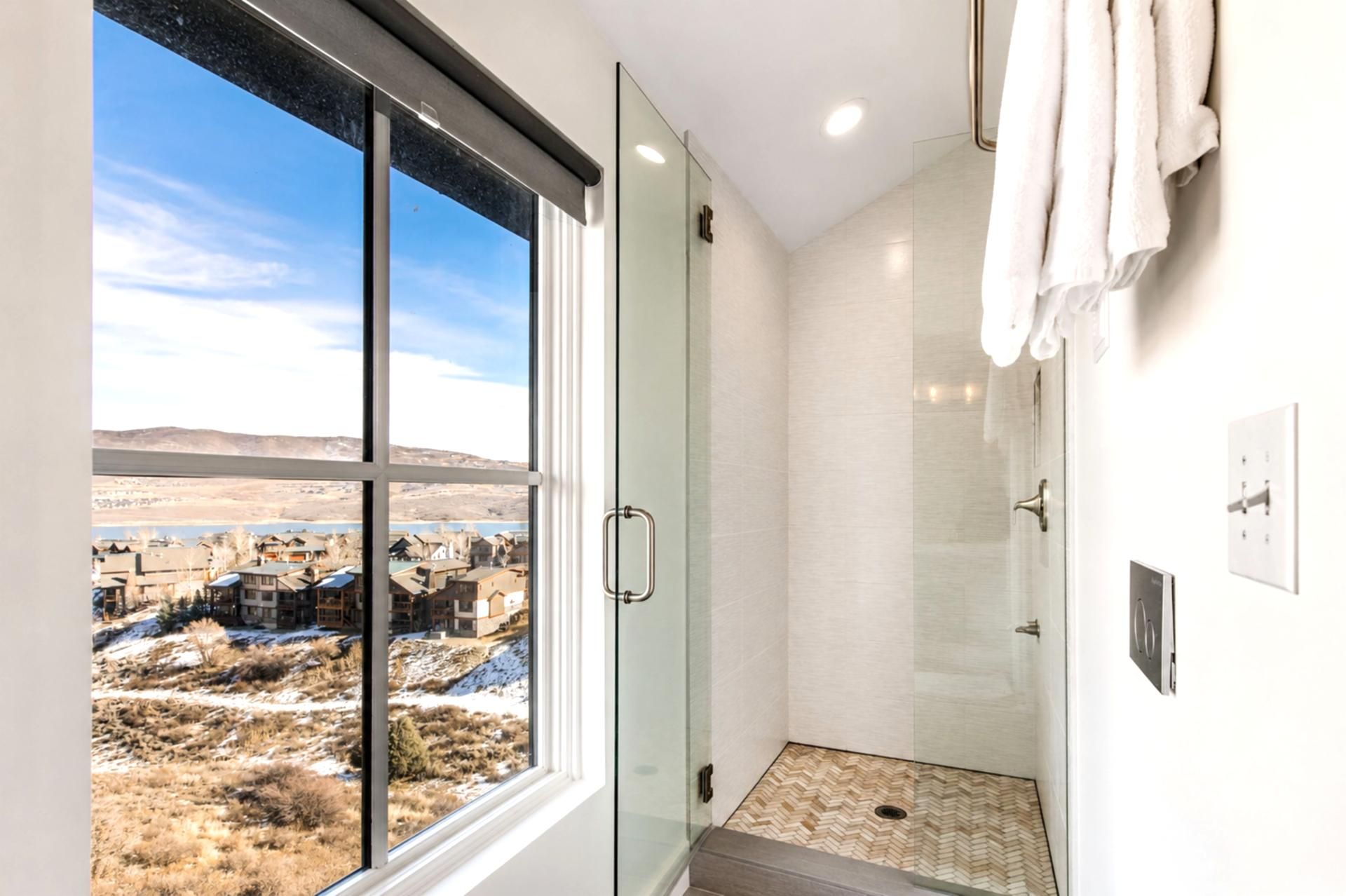 A bathroom with a glass door, a shower, and a view of a town through a large window.