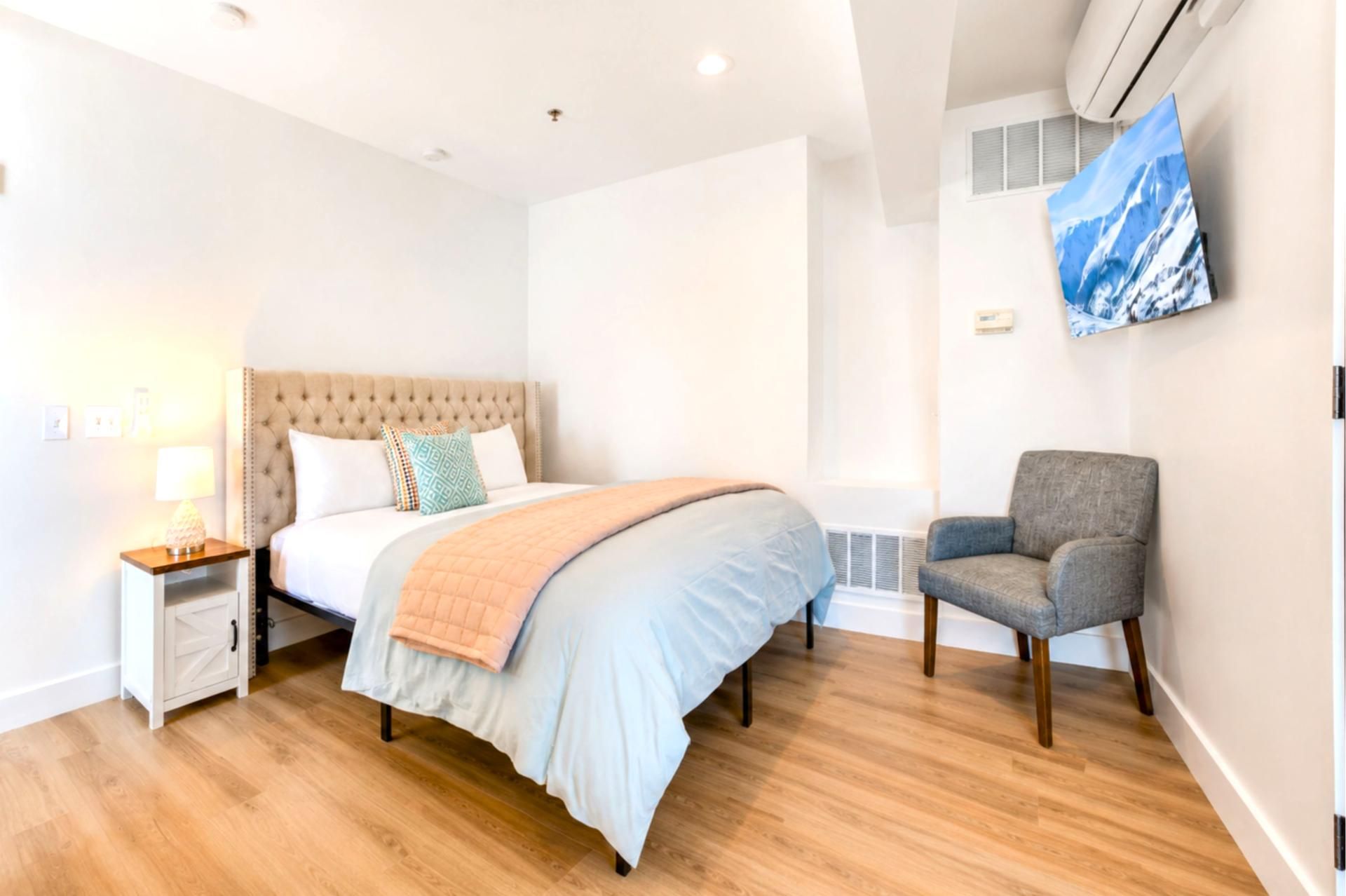 A hotel room with a bed, a chair, a nightstand, and a wall-mounted air conditioner.