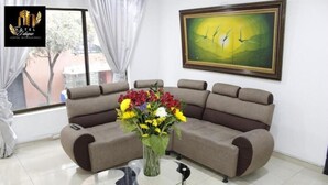 Una sala moderna con un sofá seccional color beige, una mesa de centro de vidrio y un florero con flores. Hay un cuadro en la pared y se ve una ventana con cortinas.