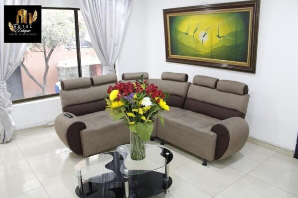 Un salon moderne avec un canapé d’angle beige, une table basse en verre et un vase de fleurs. Un tableau est accroché au mur, et une fenêtre avec des rideaux est visible.