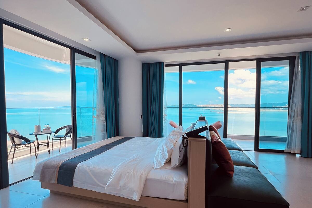 A modern hotel room with a large bed, a sofa, and a balcony with a view of the sea.