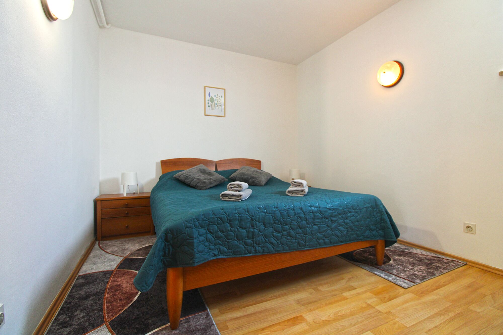 A neatly made bed with a teal bedspread and two pillows, a wooden nightstand with a lamp, and a small wall-mounted light fixture.