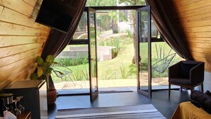 Una habitación con techos inclinados de madera, un ventanal grande, un sofá, una silla y un televisor sobre una estantería de madera.