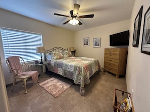 Une chambre à coucher comprenant un lit, une chaise, une commode, un téléviseur et un ventilateur de plafond.