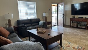 A living room with a dark leather sofa, a coffee table with a board game, a TV on the wall, and a door leading outside. - Cozy pet friendly house with a great outdoor space, and fully stocked kitchen.   (Las Cruces)