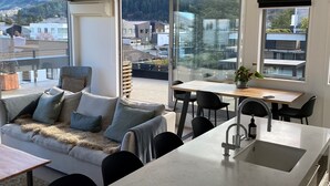 Cozinha moderna com uma ilha grande, área de jantar com mesa e cadeiras, sofá com almofadas e janelas amplas com vista para prédios e árvores.