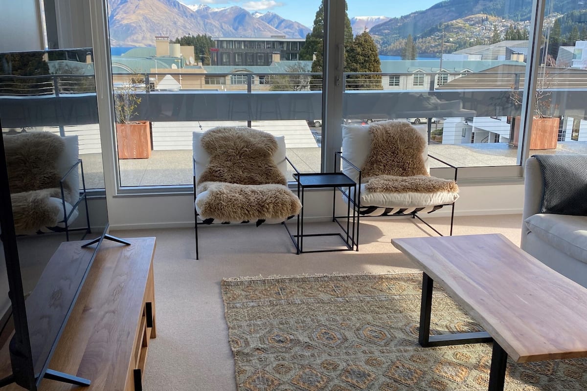 A modern living room with a large window overlooking a mountainous landscape. There are two sofas with fluffy cushions, a wooden bench, and a small table.