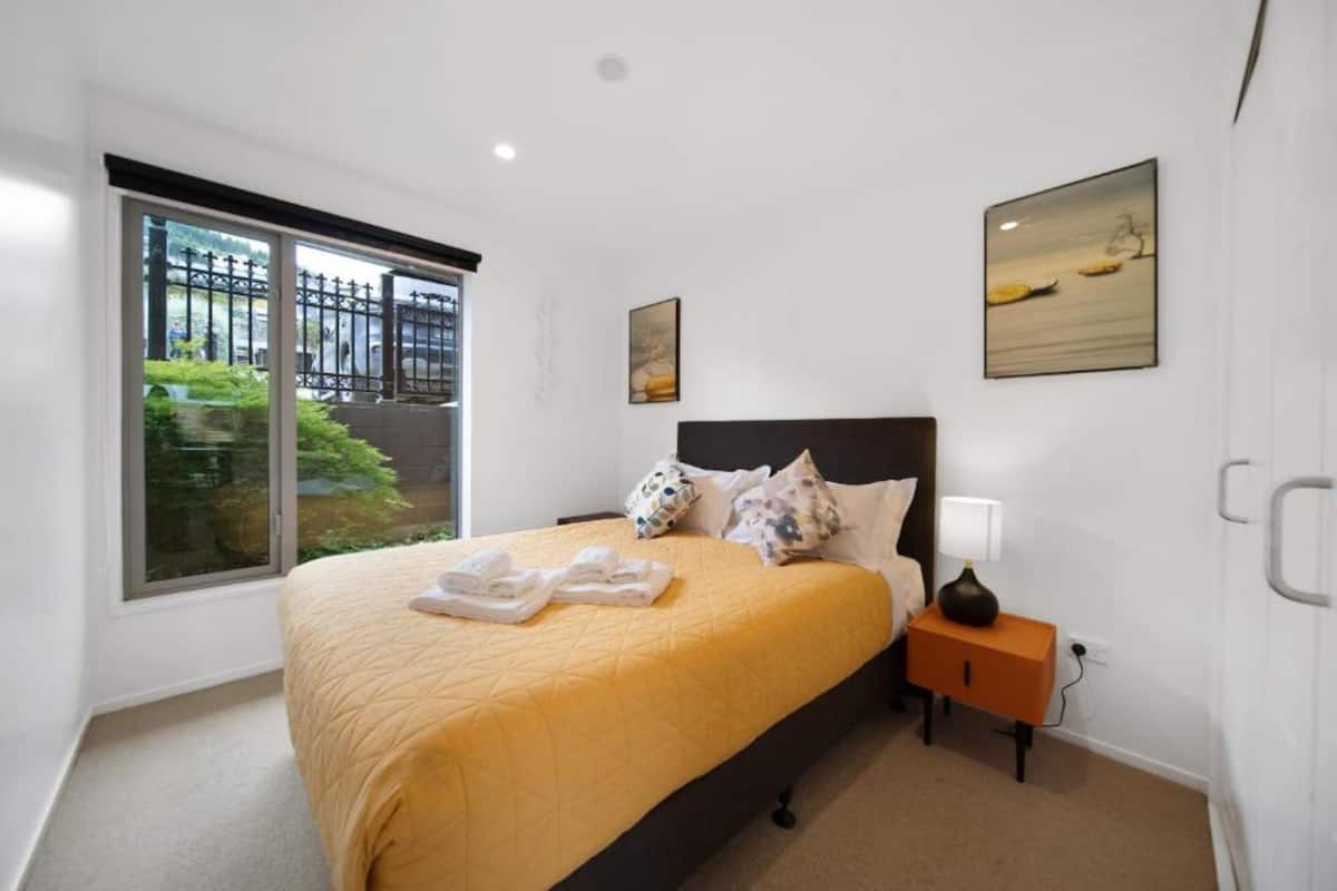 A bedroom with a large bed, a nightstand, a window with a view of greenery and a fence, and two wall-mounted paintings.