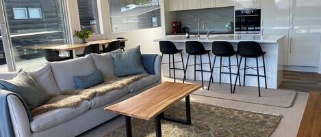 A modern living room with a sofa, coffee table, and patterned rug. A kitchen with white cabinets and black bar stools is visible in the background.