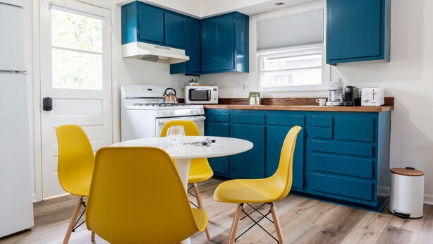 Een kleine keuken met blauwe kastjes, een witte kookplaat en gele stoelen.