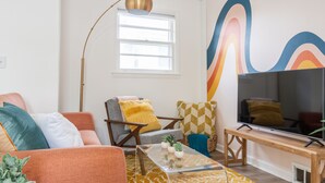 Een woonkamer met een bank, fauteuil en salontafel, een tv op een houten dressoir, en een raam met vloerlamp.