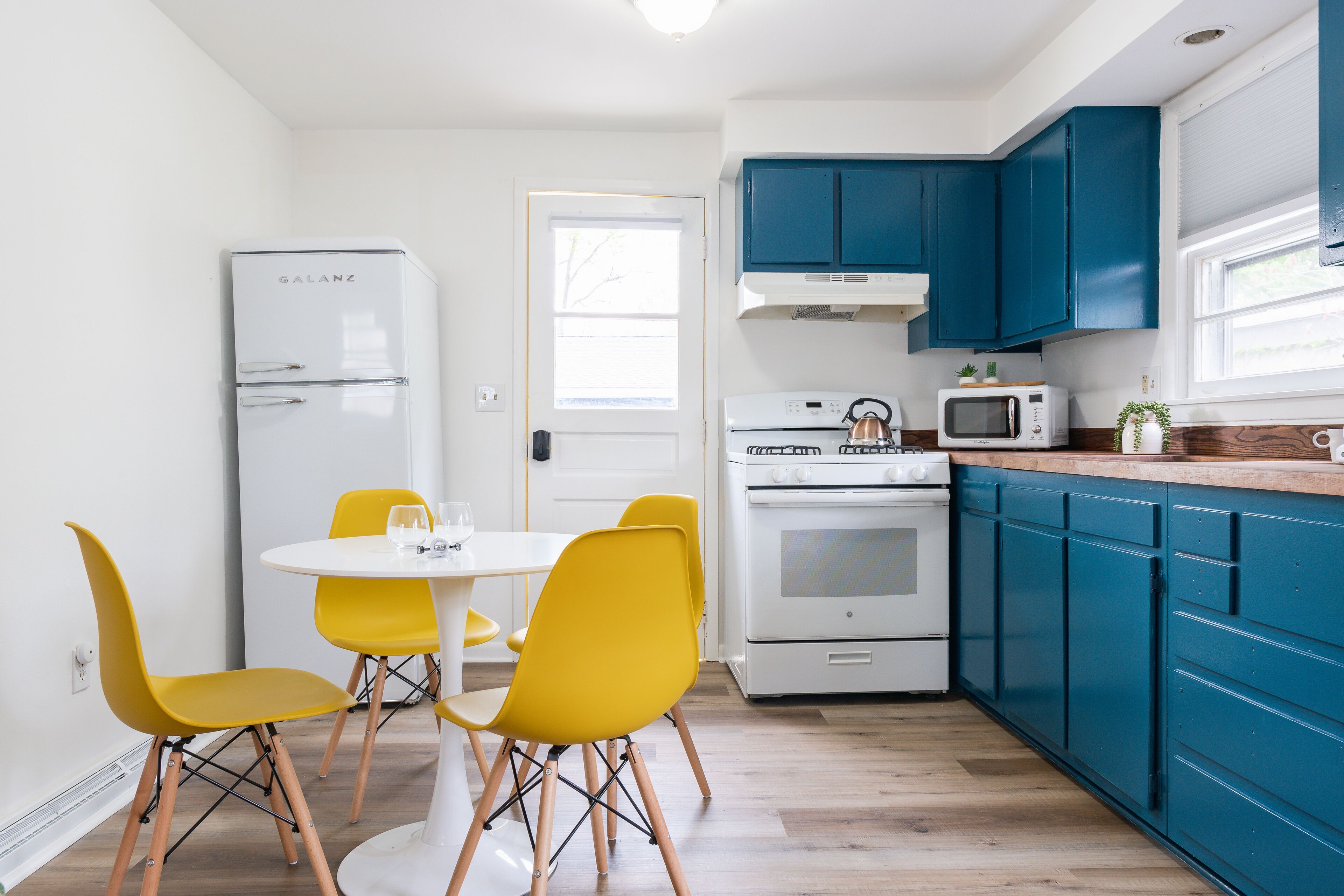 Una cocina compacta con un refrigerador blanco, un microondas y una estufa. Hay sillas amarillas alrededor de una pequeña mesa blanca. Los gabinetes azules recubren la cocina.