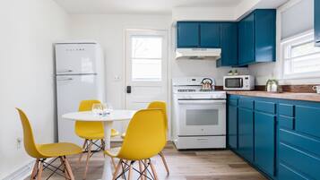 Una cocina compacta con un refrigerador blanco, un microondas y una estufa. Hay sillas amarillas alrededor de una pequeña mesa blanca. Los gabinetes azules recubren la cocina.