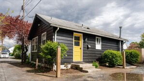 Een klein, gelijkvloers huis met een gele voordeur, een veranda en een kleine voortuin met een boom en een struik.