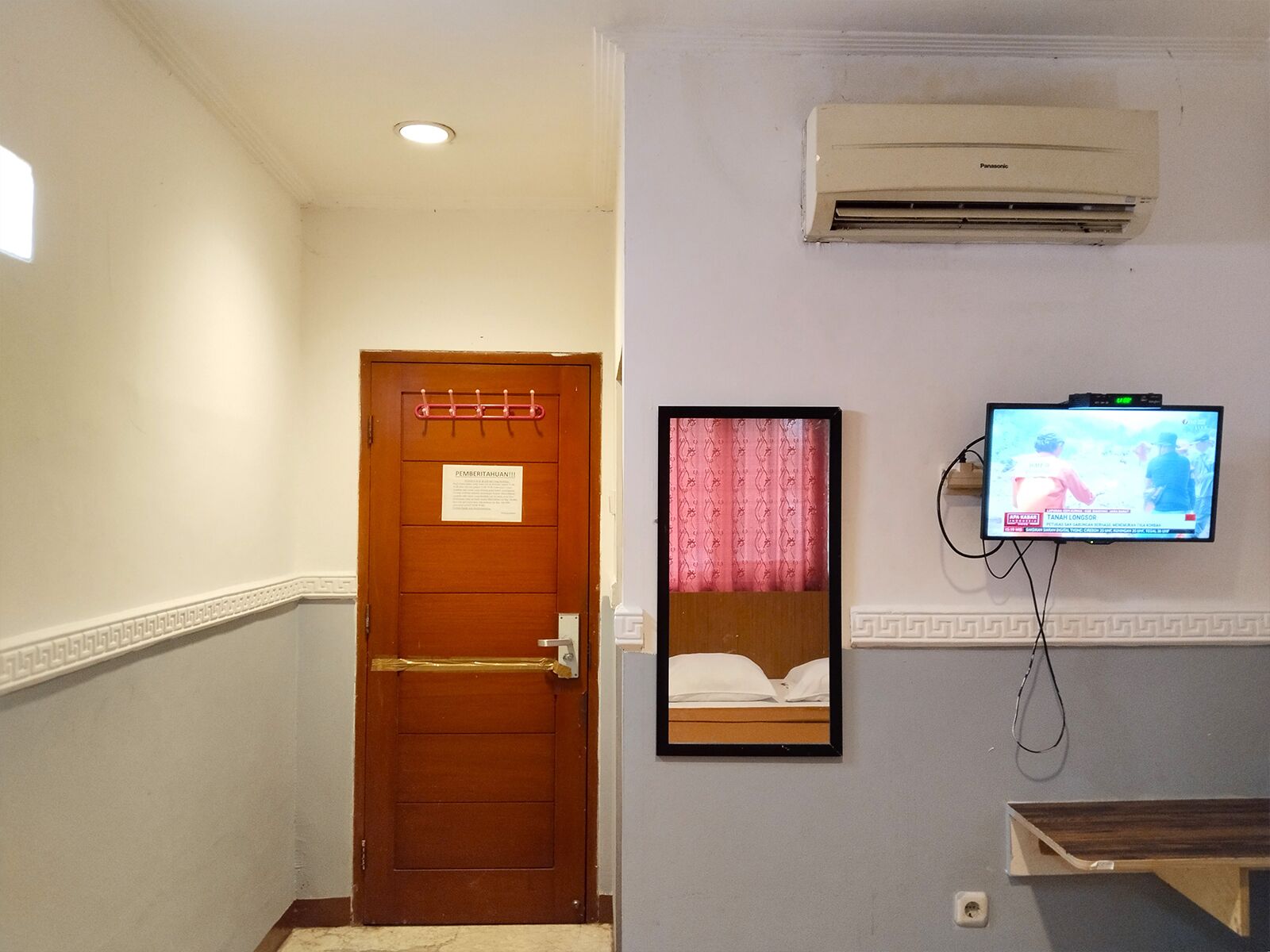Una habitación de hotel con una puerta de madera, un aire acondicionado instalado, un televisor mostrando un noticiero y un cuadro enmarcado con una cama.