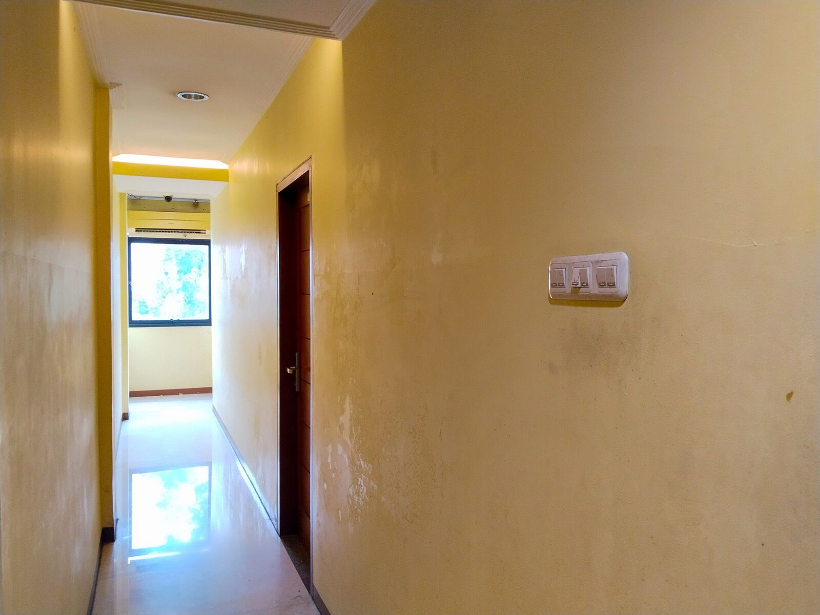 A hallway with a door on the right, a light switch, and a window at the end.