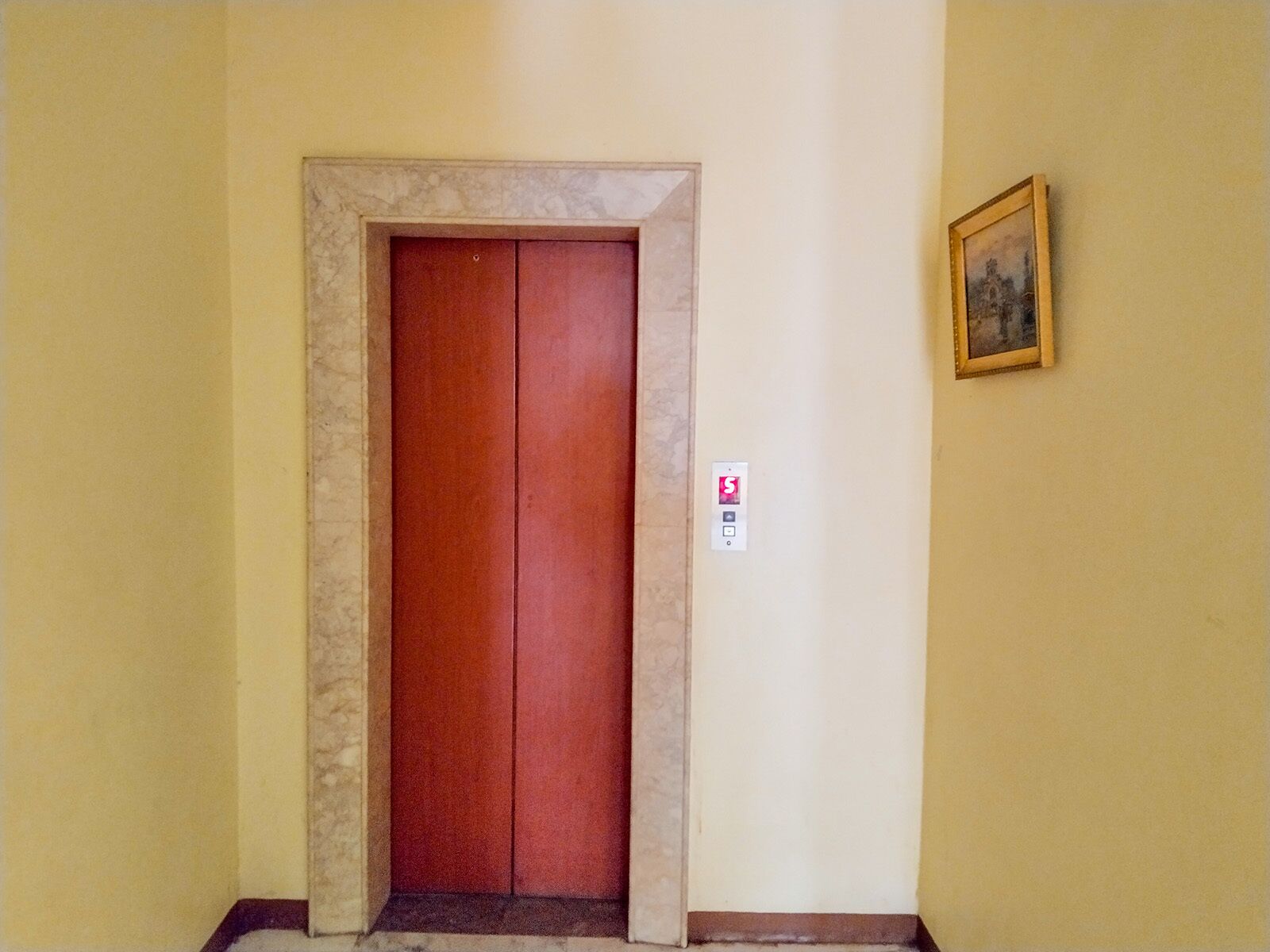 A closed elevator door with a marble frame, a fire alarm button, and a framed picture on the wall.