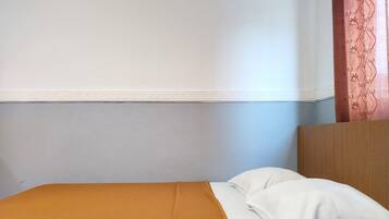 A single bed with a white pillow and a mustard-colored blanket in a room with a plain wall and a wooden headboard.