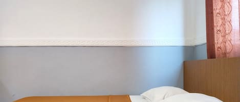 A single bed with a white pillow and a mustard-colored blanket in a room with a plain wall and a wooden headboard.
