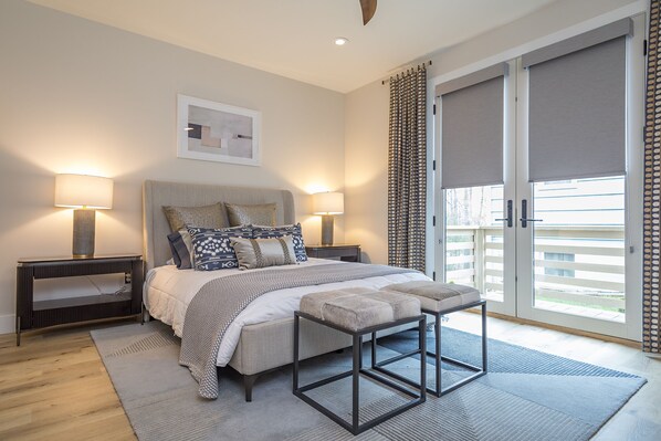 A bedroom with a large bed, two bedside tables with lamps, a bench, a rug, and a view through a sliding door.
