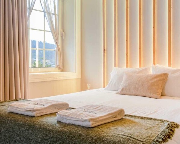 Ein Hotelzimmer mit einem großen Bett, zwei gefalteten Handtüchern, einem Fenster mit Vorhängen und Blick ins Freie.