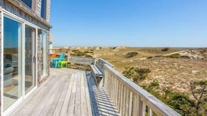 A wooden deck with a glass door leading to a room, overlooking a beach with dunes and a clear sky.