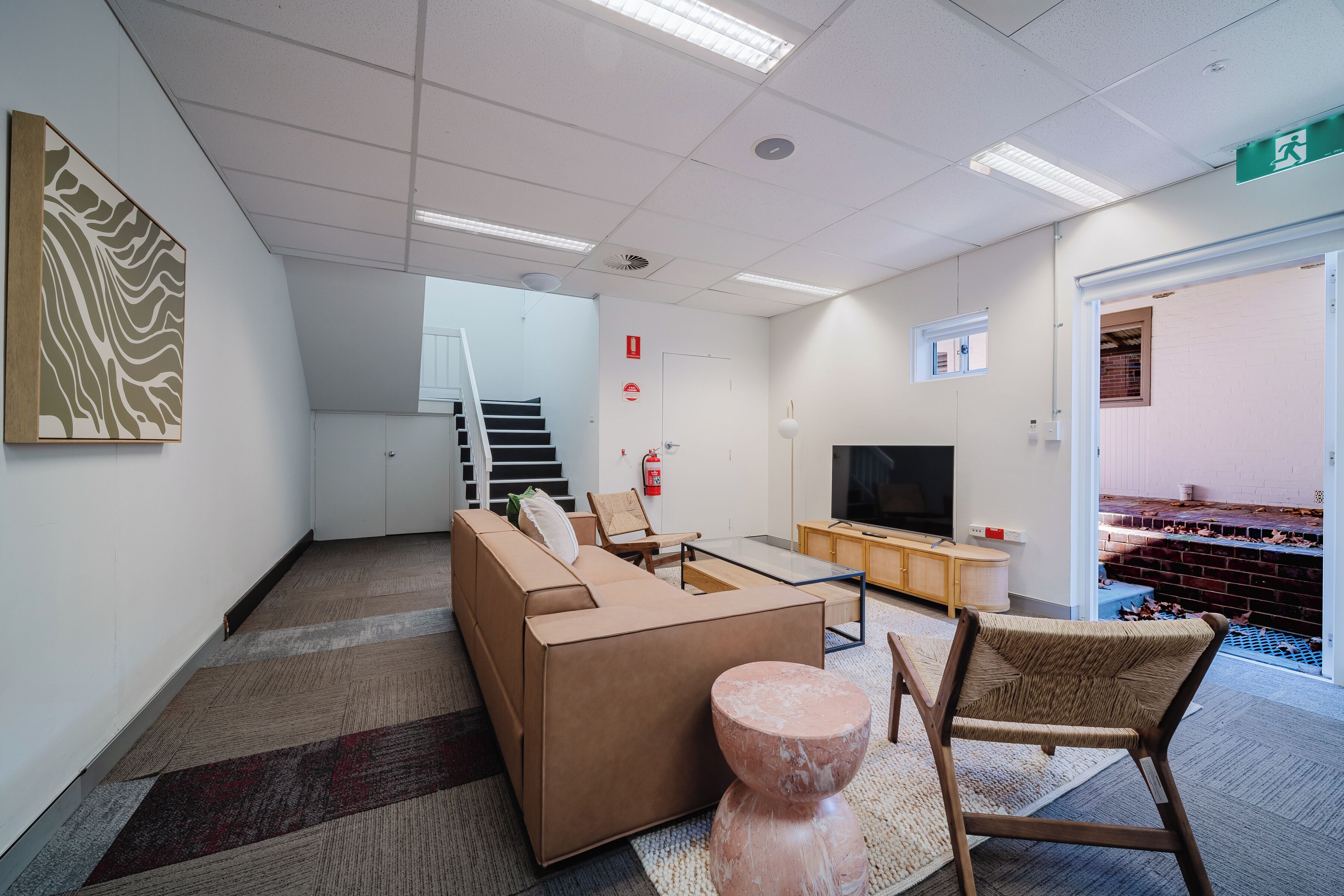 A modern living room with a sofa, coffee table, and a TV. There is a staircase leading to an upper level, a fire extinguisher, and a door with an exit sign.