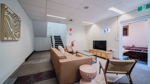 Een moderne woonkamer met een bank, salontafel en een televisie. Er is een trap die naar een hoger niveau leidt, een brandblusser en een deur met een nooduitgangbord.