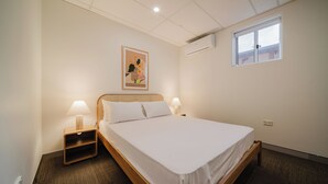 Een eenpersoonsbed met witte lakens, een nachtkastje met lamp, een wandairconditioner en een raam met uitzicht.