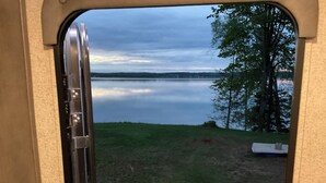 A view from a window with a metal frame, overlooking a calm lake and grassy area with trees. - Cozy couples camper with your own private 100ft lake front.   (McMillan)