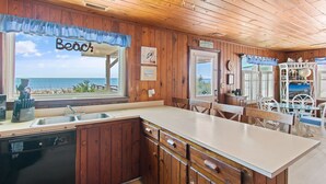 A kitchen with wooden cabinets, a large window with a view of the beach, and a dining area with white chairs. - "Lifesaver" is a 2 Bedroom, 1 Bathroom, Oceanfront Home! (Holden Beach)