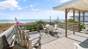 A wooden deck with a view of the beach, a covered seating area, and a flag. - "Lifesaver" is a 2 Bedroom, 1 Bathroom, Oceanfront Home! (Holden Beach)