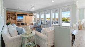 A spacious living room with a white sofa, a small round table, and a television. There is a wooden entertainment unit and a large window with a view of the outdoors.