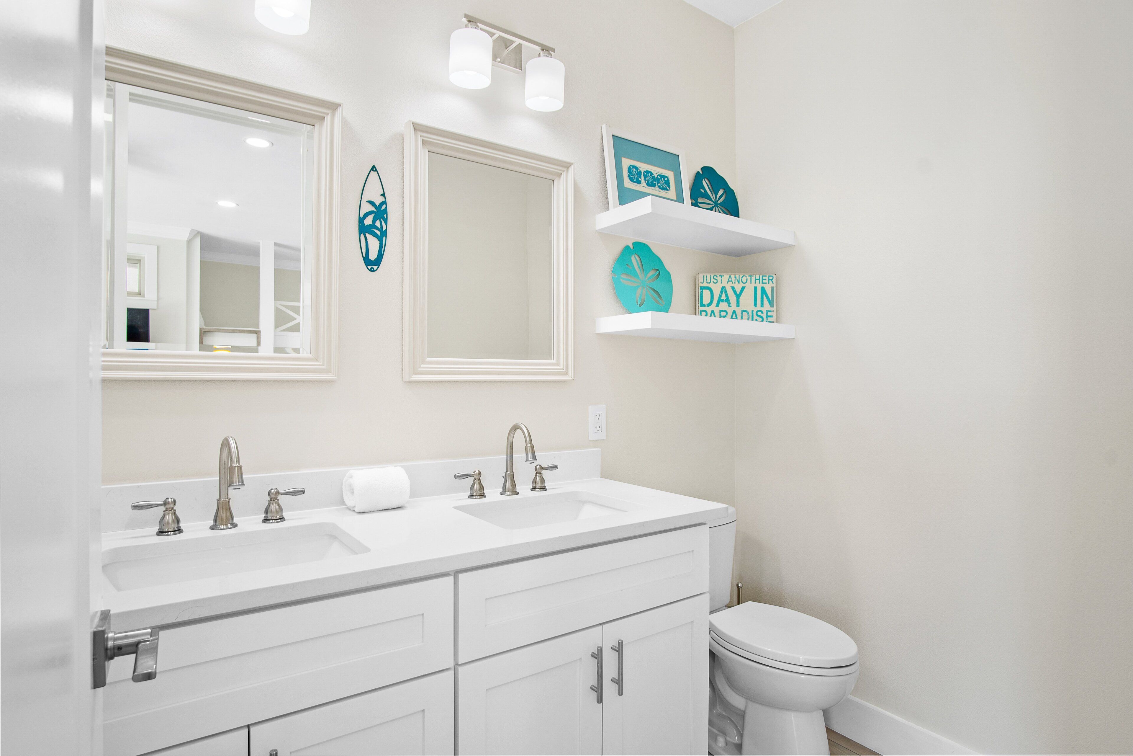 A bathroom with a white vanity, two sinks, a toilet, and decorative wall shelves.
