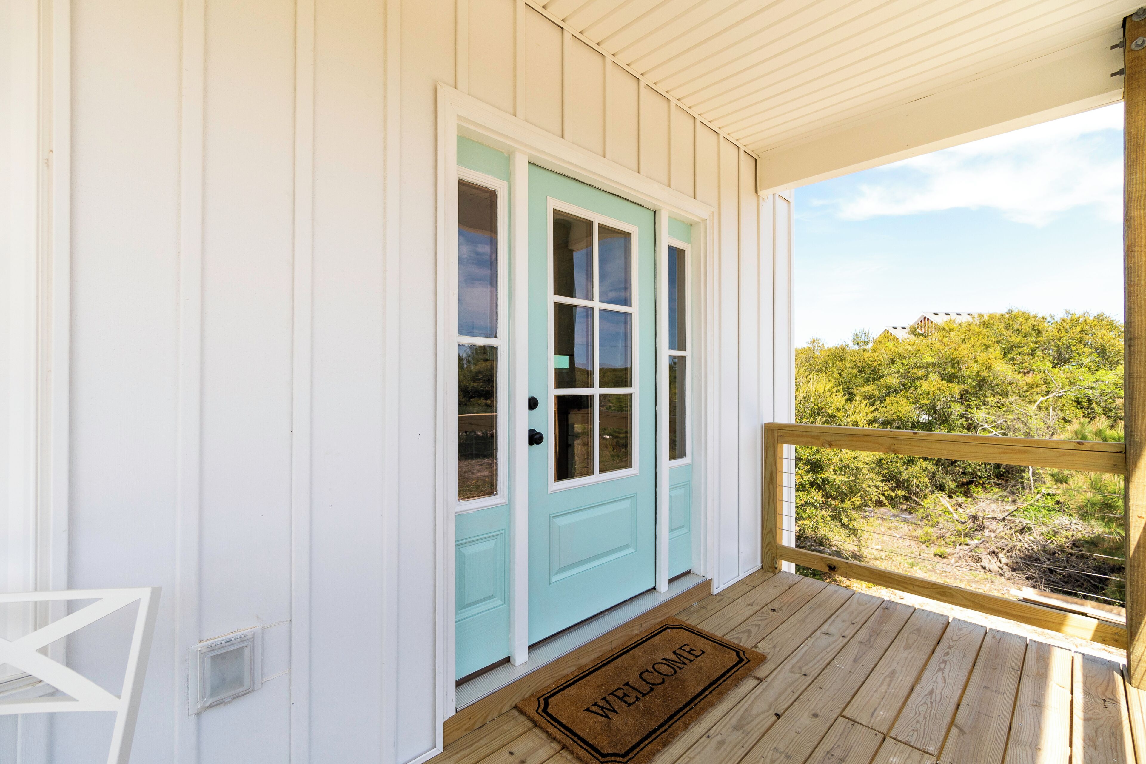 A wooden porch with a welcome mat, a light blue door, and a view of greenery.