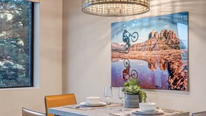 A dining area with a table set for two, a large framed picture on the wall, and a window with a view of trees.