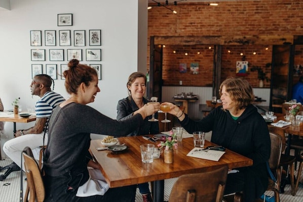 Um grupo de pessoas jantando em uma mesa de madeira em um restaurante, com uma parede de tijolos e obras de arte emolduradas ao fundo.