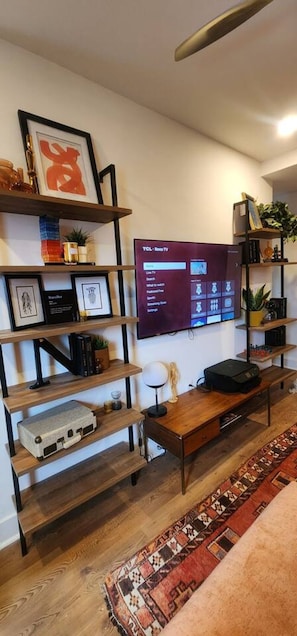 Uma sala de estar com televisão de tela plana na parede, prateleiras de madeira com diversos objetos, uma mesa de centro e um tapete estampado.