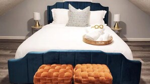 A bedroom with a vaulted ceiling, a bed with a dark blue headboard, white bedding, two matching orange ottomans, and bedside lamps.