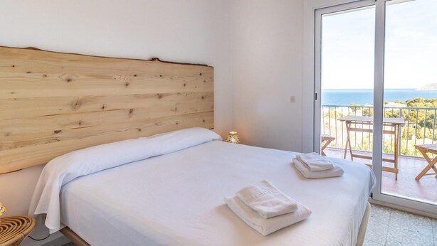 A bedroom with a wooden headboard, a large bed with white linens, two folded towels, a small table with a lamp, and a balcony with a view of the sea.