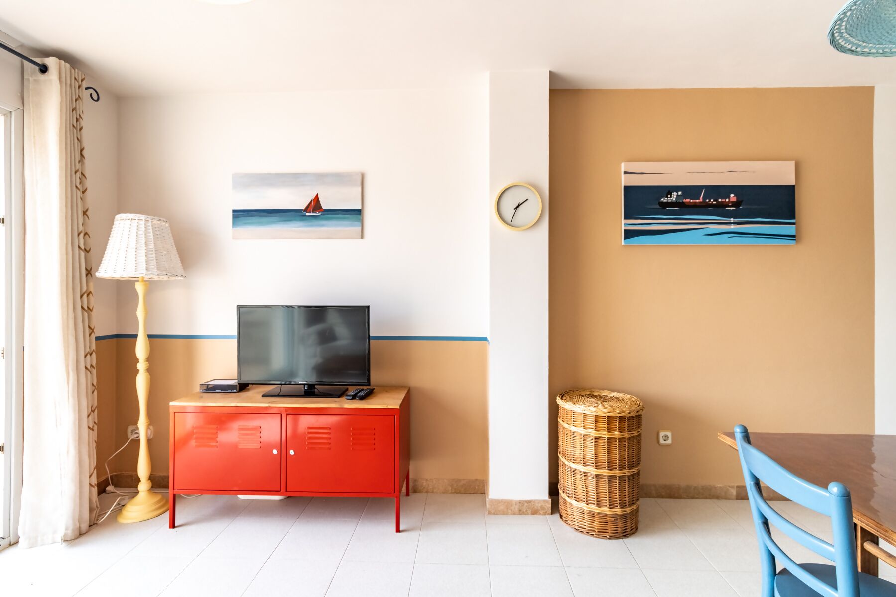 Una habitación con televisión sobre un mueble rojo, una cesta de mimbre, un reloj y dos cuadros en la pared.