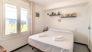 A bedroom with a bed, a nightstand, a window with curtains, a view of the outside, and a shelf with books and decorative items. - Cap Ras 04.3 - Apartment with terrace and sea view (Llança)