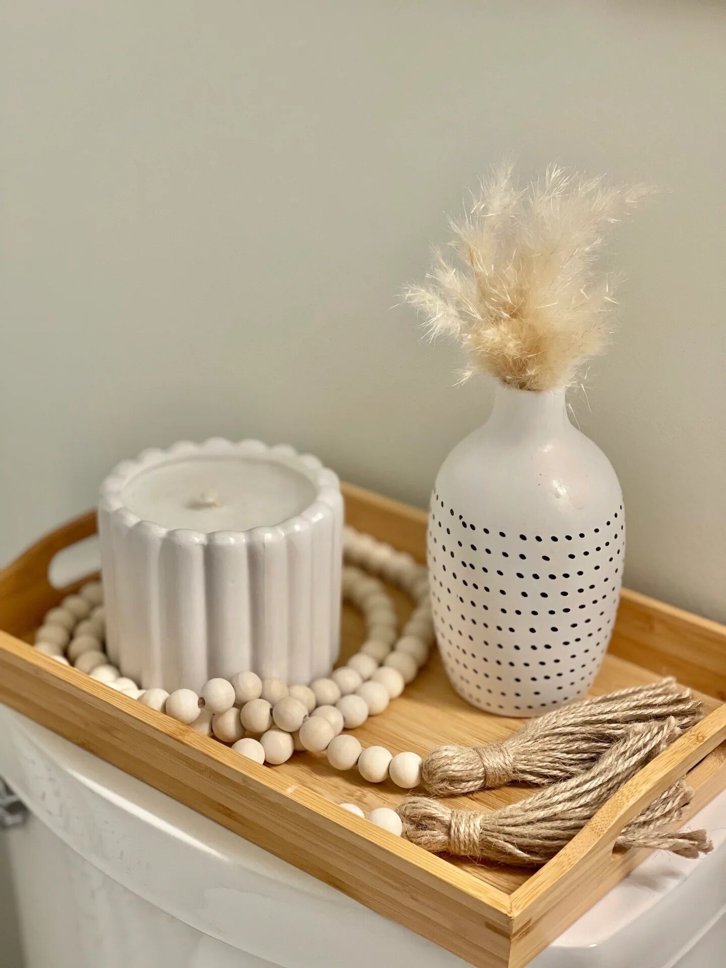 A white ceramic candle holder with a textured design, a white vase with a feather decoration, and a wooden tray with white beads and twine.