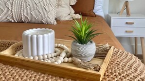 Een netjes opgemaakt bed met een houten dienblad waarop een witte kaars, een kleine potplant en een draadje witte kralen staan.