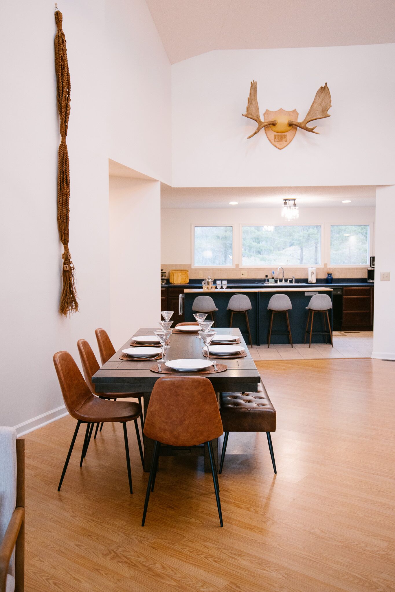 A modern dining area with a wooden table set for six, surrounded by chairs. A kitchen with a large island and bar stools is visible in the background.