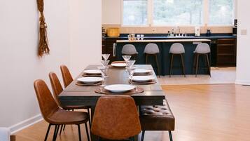 A modern dining area with a wooden table set for six, surrounded by chairs. A kitchen with a large island and bar stools is visible in the background.