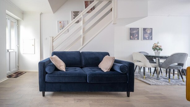 Ein modernes Wohnzimmer mit einem dunkelblauen Sofa, einer Treppe, einem Essbereich mit Tisch und Stühlen sowie einer Tür links.