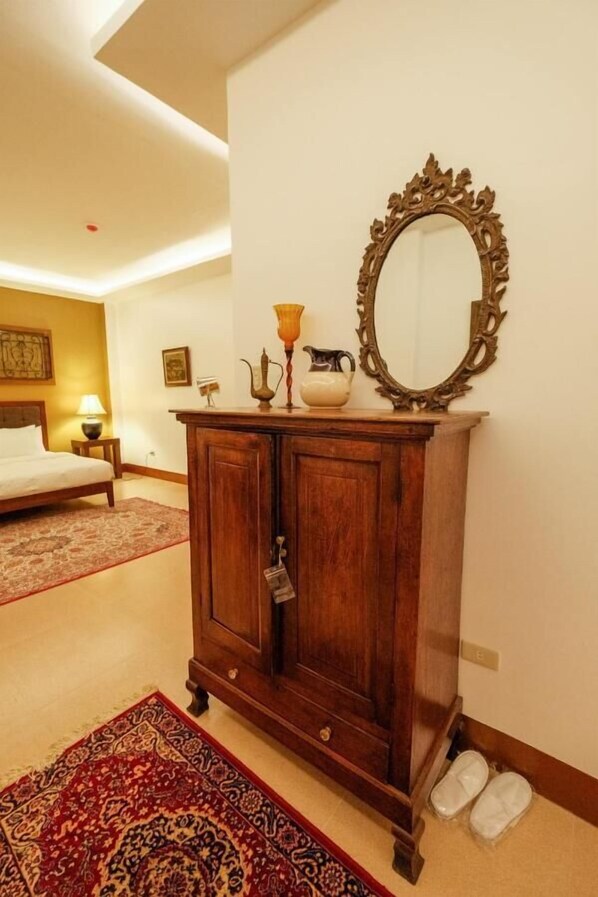 Une chambre d’hôtel avec une armoire en bois, un grand miroir, un lit, un tapis rouge à motifs et une paire de chaussons posés sur le sol.